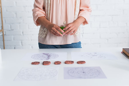 partial view of fortune teller holding vial of essential oil near clay runes and astrological mapsの写真素材