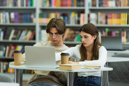 teenage girl and guy in eyeglasses studying near laptop and books in library