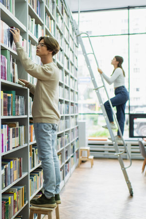 student in eyeglasses choosing books in library near teenage girl on ladderの写真素材
