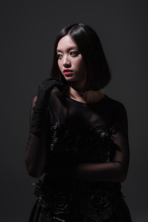 brunette asian model in black gothic outfit and gloves isolated on dark grey