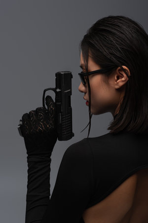 side view of dangerous asian woman in total black outfit and stylish sunglasses holding gun isolated...