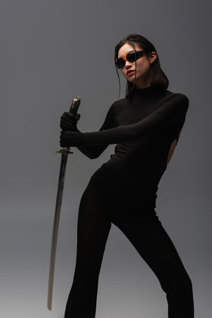 brunette asian woman in sunglasses and black clothes holding katana isolated on grey