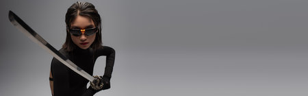 brunette asian woman in sunglasses and black outfit holding katana isolated on grey, banner
