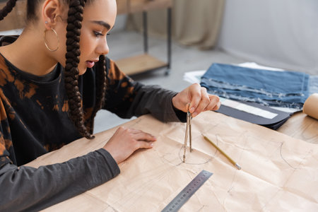 African american designer drawing with compass on sewing print near blurred cloth in workshop