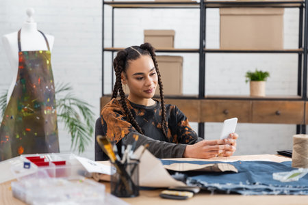 African american designer using smartphone near blurred sewing equipment in workshopの写真素材