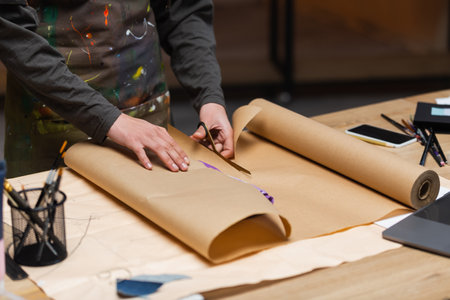 Cropped view of african american craftswoman cutting craft paper while packaging clothes in workshop