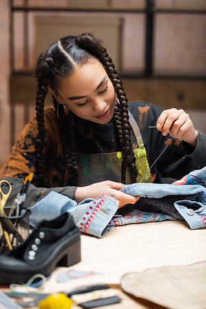 Young african american craftswoman embroidering denim jacket in workshopの写真素材