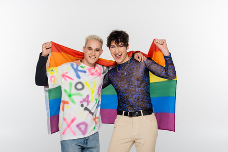 excited queer person and happy gay man holding lgbt flag and looking at camera isolated on greyの写真素材
