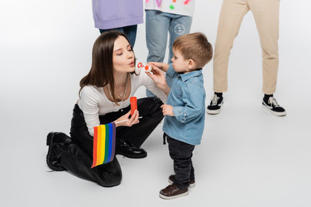 young lesbian woman with lgbtq flag blowing soap bubbles near toddler boy on grey backgroundの写真素材