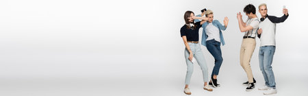 young woman showing victory sign and dancing near friends taking photo on smartphones on grey...