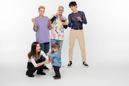 happy lgbtq friends blowing soap bubbles near toddler boy on grey background