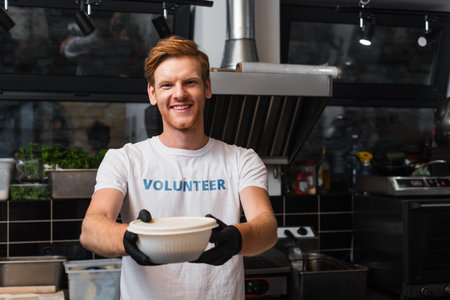 happy redhead volunteer offering plastic bowl in kitchenの写真素材