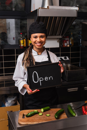 happy african american chef holding chalkboard with open lettering in kitchen