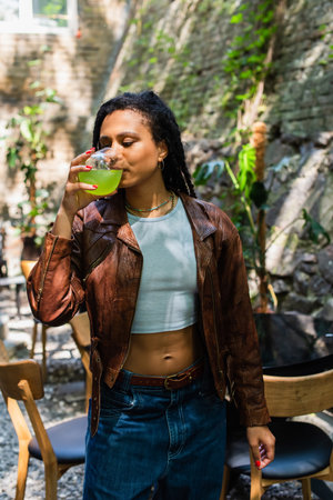 young african american woman in leather jacket drinking green cocktail outside