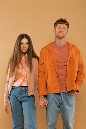 happy young couple in autumnal outfits holding hands while standing isolated on beige