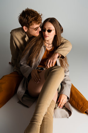 stylish young man in sunglasses hugging girlfriend and sitting on greyの写真素材