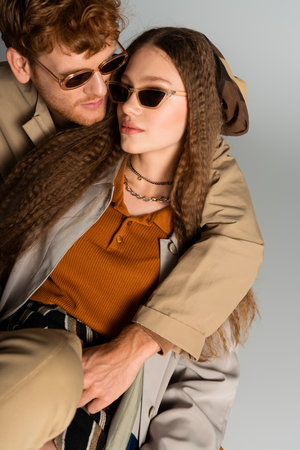 stylish young man in trendy sunglasses hugging girlfriend isolated on greyの写真素材
