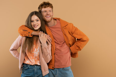 young happy man with red hair looking at camera and hugging pretty girlfriend isolated on beigeの写真素材