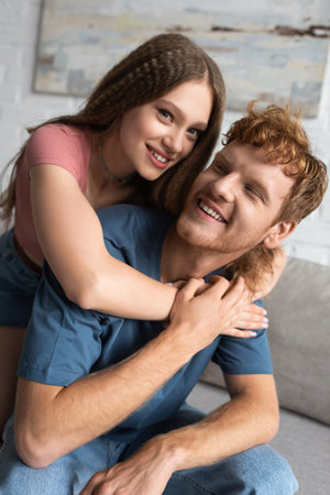 happy teen girl smiling and hugging redhead boyfriend in living roomの写真素材