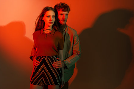 young redhead man standing and hugging hips of stylish girl in skirt on red background