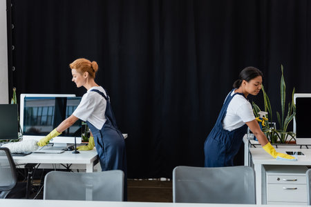 side view of multiethnic women cleaning office near computer monitors with blank screenの写真素材