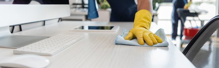 cropped view of cleaner in rubber glove washing desk near computer monitor and digital tablet,...
