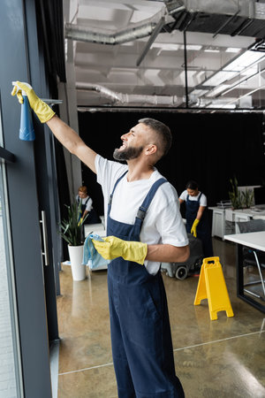 bearded man in workwear cleaning window with detergent near multiethnic women working in office