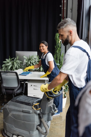 bi-racial woman smiling at camera near man with floor scrubber machine on blurred foregroundの写真素材