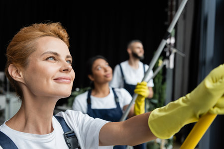 selective focus of happy woman washing windows near blurred multiethnic cleaning teamの写真素材