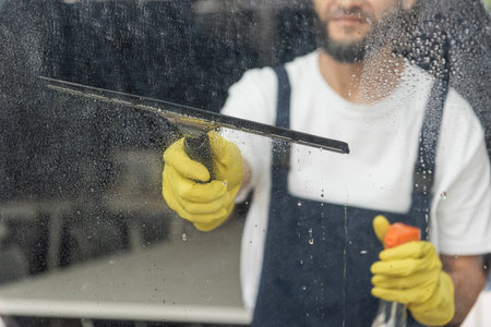 cropped view of man with detergent and window wiper washing glass in officeの写真素材