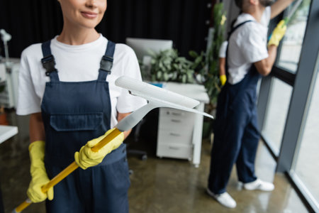 cropped view of blurred woman in overalls holding window squeegee near team working on blurred...