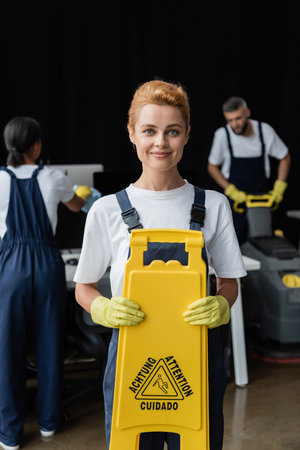 cheerful woman with caution sign board looking at camera while colleagues working on blurred...