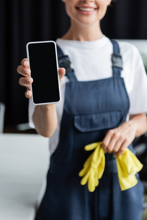 partial view of blurred woman in overalls holding rubber gloves and smartphone with blank screenの写真素材