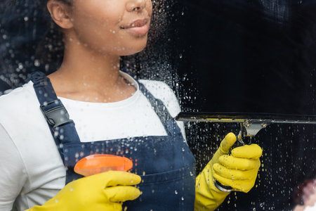 cropped view of bi-racial woman with detergent and window squeegee near wet glass in officeの写真素材