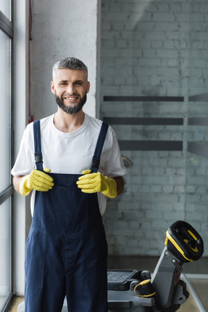 cheerful man in rubber gloves and overalls smiling at camera near electrical floor scrubber machineの写真素材