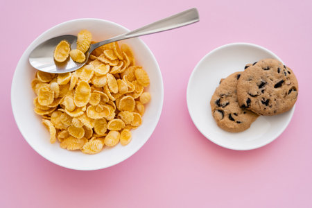 top view of bowl with tasty corn flakes and spoon near saucer with chocolate chip cookies on pinkの素材 [FY310191620684]