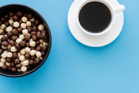 top view of bowl with tasty cereal balls near cup of black coffee isolated on blueの写真素材