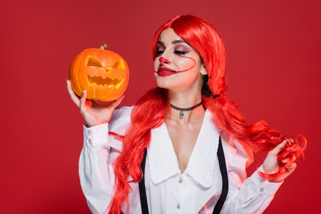 smiling woman with bright hair and clown makeup holding carved pumpkin isolated on redの写真素材