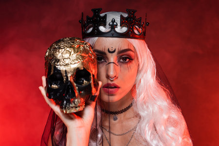blonde woman with witch makeup wearing black crown and veil while looking at camera near golden skull on red backgroundの写真素材