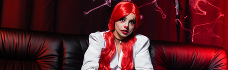 redhead woman with ponytails and halloween makeup on black couch and dark background with spiderweb,...