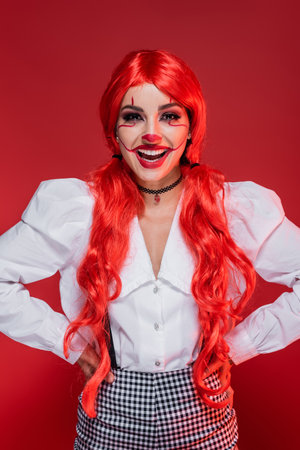 smiling woman with clown makeup and bright hair standing with hands on hips isolated on redの写真素材