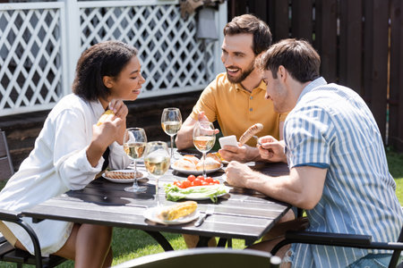 Smiling man pointing at smartphone near interracial friends and food in backyardの写真素材