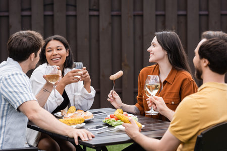 cheerful multiethnic friends holding glasses with wine and having meal during bbq party in backyardの写真素材