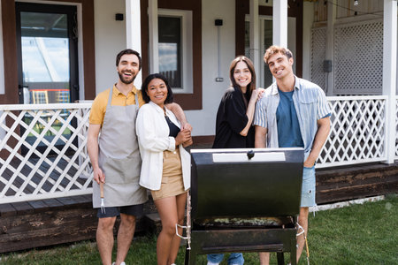 Positive interracial friends hugging and looking at camera near grill in backyard