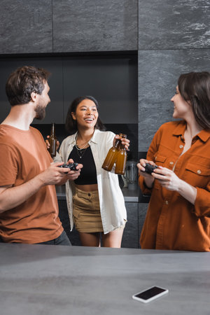 cheerful bi-racial woman giving beer to friends with joysticks in kitchenの写真素材