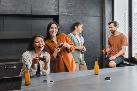 happy interracial women playing video game near friends in kitchenの写真素材