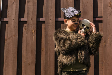 happy asian boy in halloween werewolf costume embracing spooky skull near wooden fenceの写真素材
