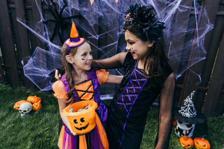 Smiling girls with halloween makeup holding candies in bucket while standing near decor in backyardの写真素材