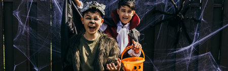 Asian children in halloween costumes holding bucket with candies near spider web on fence in...