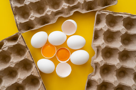 Top view of eggs and raw yolks near carton trays on yellow background